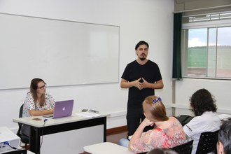 Sala de aula. Uma mulher está sentada na mesa do professor. Um homem está de pé ao lado direito da mesa e duas mulheres aparecem sentadas de costas.